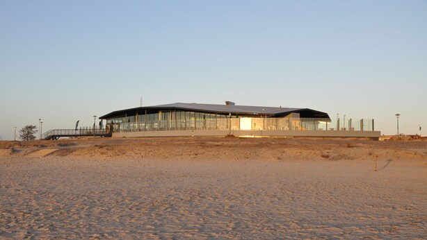 A modern building with a glass facade and a flat roof, situated on a sandy terrain.