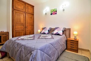 Une chambre avec une grande armoire en bois, un lit recouvert d’une courtepointe violette, deux lampes de chevet et une petite table de nuit en bois.