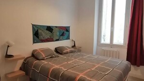A bedroom with a bed, a nightstand, a wall-mounted lamp, a window with curtains, and a painting on the wall. - Newly renovated apartment Joliette Marseille (Marseille)