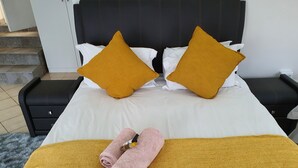 Een net opgemaakt bed met witte en mosterdkleurige kussens, een gele plaid en roze handdoeken.