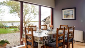 Uma área de refeições com mesa posta para quatro pessoas, grandes janelas com vista para um lago e árvores, e uma moldura com uma imagem na parede.