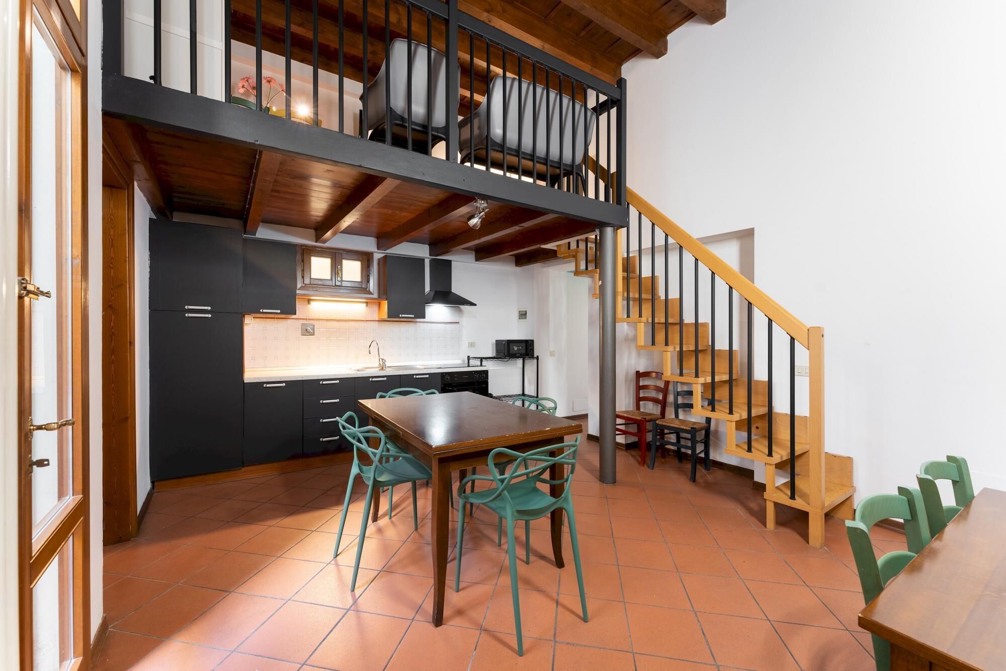A kitchen with a dining area, featuring a wooden staircase, black cabinets, and green chairs.