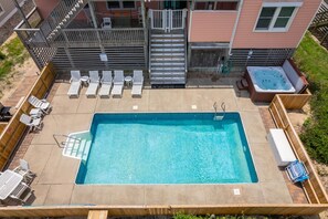 Una piscina rectangular con un muelle de madera alrededor, un jacuzzi a la derecha y un juego de escaleras que llevan a un edificio.
