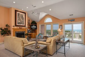 A living room with a fireplace, a sofa, a coffee table, and a balcony with a view of the ocean. - Barefootin'- Oceanfront, private pool, hot tub, pool table, private walkway to the beach! (Nags Head)