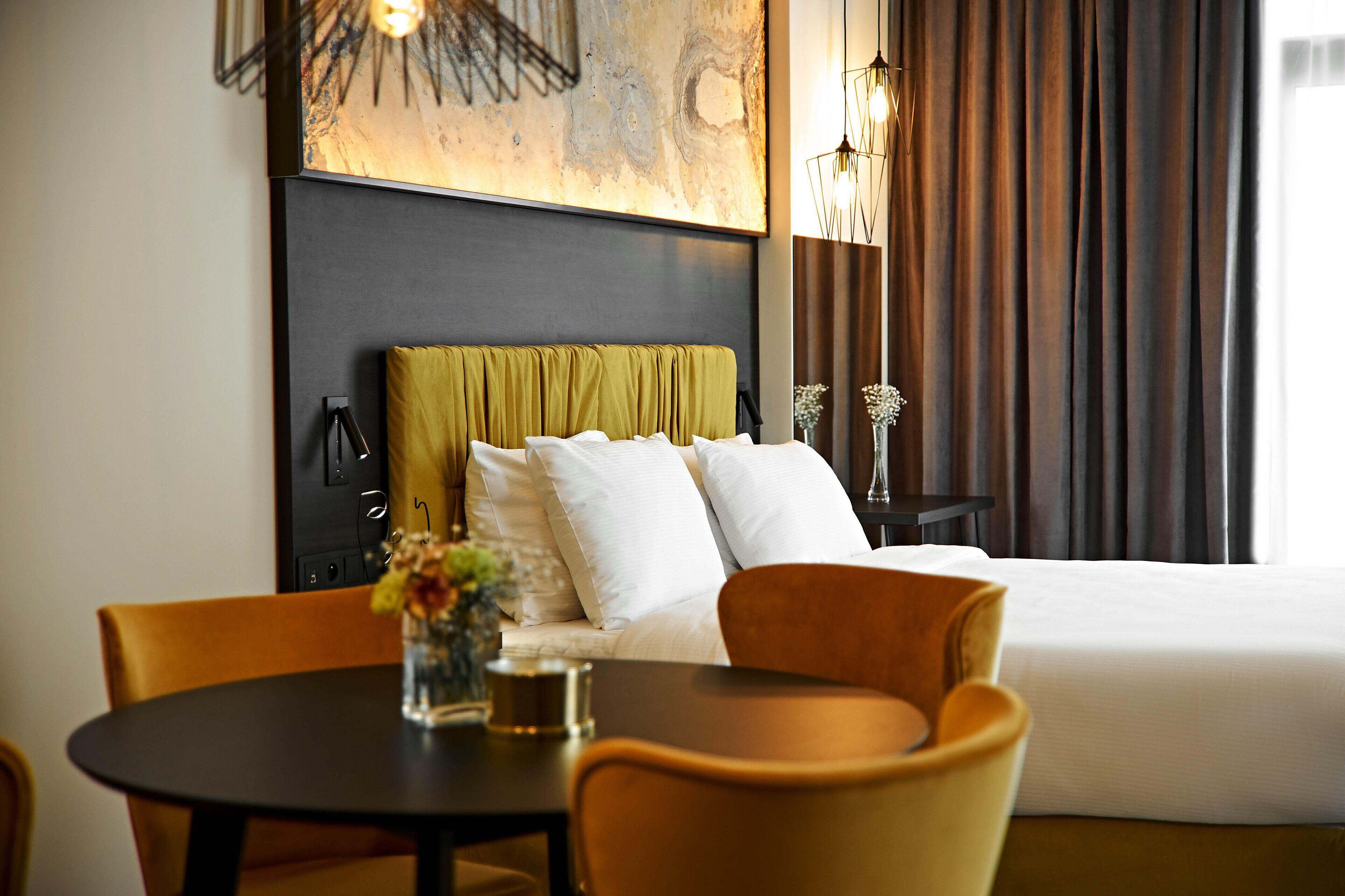 Habitación de hotel con cama, una mesa de comedor con sillas, un florero con flores y un cuadro en la pared.
