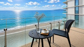 A balcony with a glass railing overlooking a beach and ocean, a table with a chair, a vase with flowers, and a cup of coffee. - Miramare Magnetic Beach Hotel (Kobuleti)
