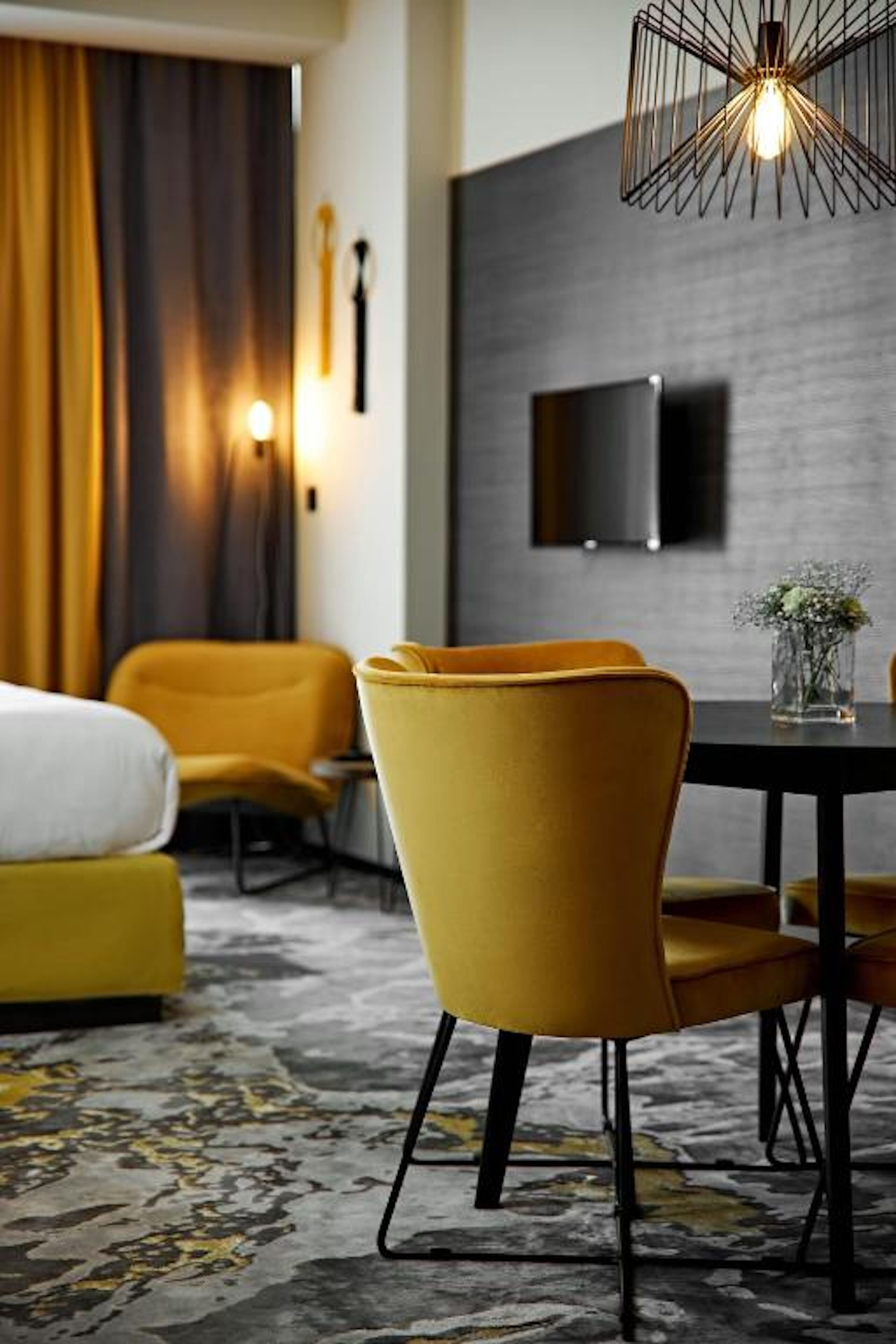 A modern hotel room with a yellow armchair, a dining table, and a TV mounted on the wall.