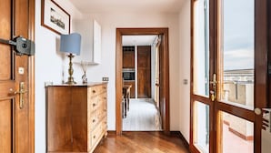 A hallway with wooden flooring, a wooden cabinet with drawers, a lamp, and a door leading to a balcony with a view. - Appartamento Portinari (Florence)