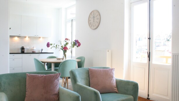 Une cuisine moderne avec des meubles blancs, une table à manger avec des chaises et une grande fenêtre donnant sur des immeubles.