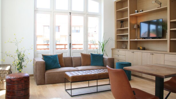 A living room with a sofa, ottoman, and a television on a wooden shelf. - De Torre 1 - luxury 1-bedroom apartment in iconic building (De Haan)