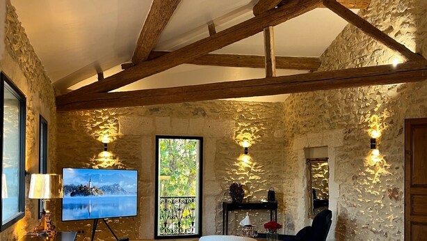 A bedroom with a bed, a desk with a lamp, a chair, and a TV displaying a scenic view. - Domaine de la Trinité (Mauguio)