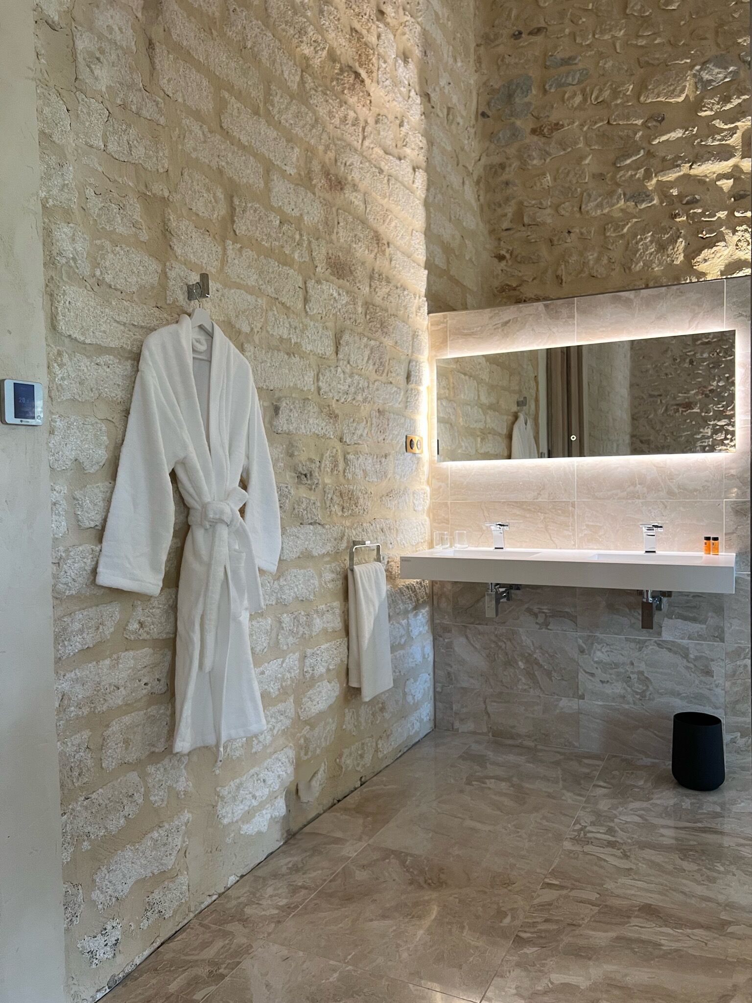 A bathroom with stone walls, a robe hanging on the wall, a mirror, and a sink with two faucets.