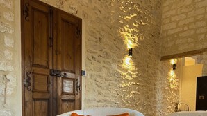 A room with a stone wall, a wooden door, a white armchair with an orange cushion, and a bed with a white bedspread. - Domaine de la Trinité (Mauguio)