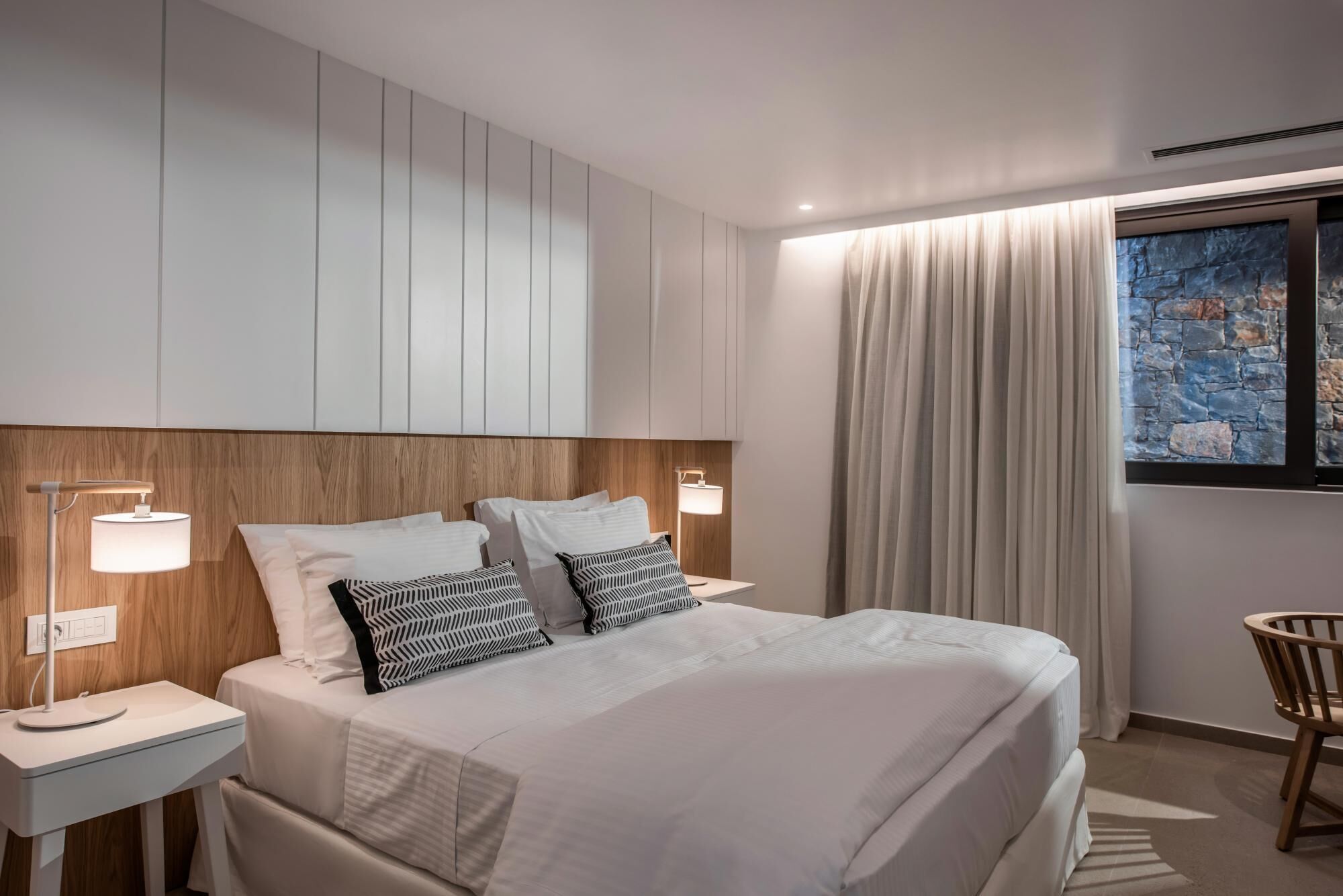 Une chambre d’hôtel moderne avec un grand lit, deux lampes de chevet, une tête de lit en bois, une œuvre d’art murale en pierre et une vue sur l’extérieur.