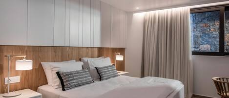 Une chambre d’hôtel moderne avec un grand lit, deux lampes de chevet, une tête de lit en bois, une œuvre d’art murale en pierre et une vue sur l’extérieur.