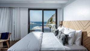 A modern hotel room with a large bed, a wooden headboard, and a balcony with a view of the sea and cliffs.