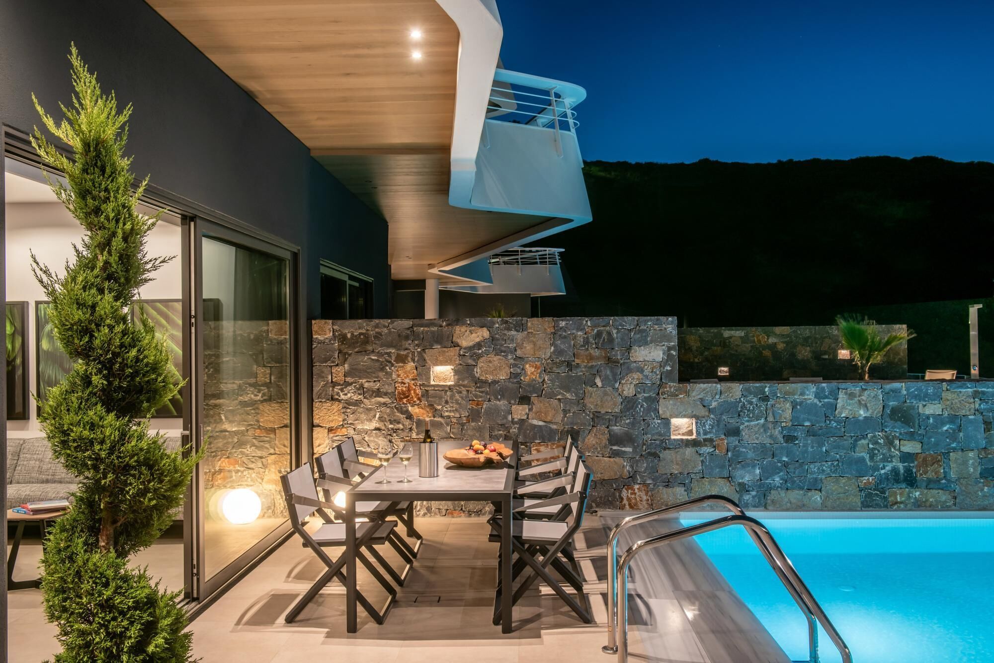An outdoor dining area with a stone wall, a pool, and a modern building with a glass door.