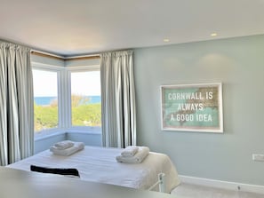 Une chambre à coucher avec un lit, deux fenêtres avec des rideaux, une citation encadrée au mur et une vue sur la mer.