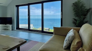 A modern living room with a large sliding glass door leading to a balcony overlooking the sea. - The Old Coastguard Lookout (Port Isaac)