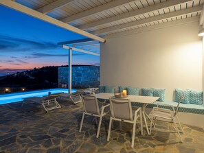 An outdoor patio with stone flooring, a table with chairs, a candle, and a view of a pool and landscape at dusk.