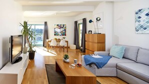 A modern living room with a flat-screen TV, a wooden coffee table, a grey sofa, and a dining area with a table and chairs.