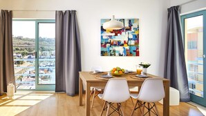 A dining area with a wooden table, white chairs, a fruit bowl, and a colorful abstract painting.