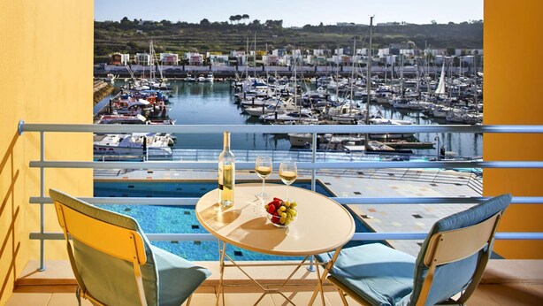Un balcon avec une table de billard, deux chaises et une table sur laquelle se trouvent une bouteille et des verres. On aperçoit une marina avec des bateaux au loin.
