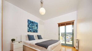 Une chambre moderne avec un grand lit, une table de chevet, une commode, une lampe sur pied, un luminaire au plafond, une fenêtre avec vue et un tableau accroché au mur.