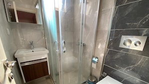 A bathroom with a glass-enclosed shower, a sink with a mirror, a toilet, and a trash bin. - Bella Rose Hotel (Fethiye)