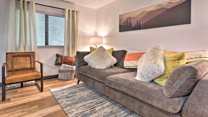 Ein Wohnzimmer mit einem braunen Ledersessel, einem grauen Sofa mit verschiedenen Kissen, einem gemusterten Teppich, einem Fenster mit Jalousien und einem an der Wand hängenden Gemälde.
