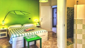 Ein Zimmer mit einem Bett, einem Nachttisch, einem grünen Hocker, einer Pendelleuchte und einem Duschbereich mit Mosaikfliesen.