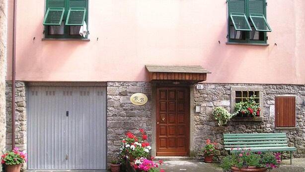 Ein Gebäude mit einer Stein-Fassade, einer Holztür und einer grauen Garagentür. Vor der Tür stehen Blumentöpfe mit Blumen und rechts daneben eine Bank.