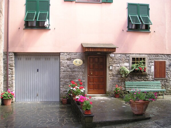 A building with a stone facade, a wooden door, and a grey garage door. There are potted plants with flowers in front of the door and a bench to the right.