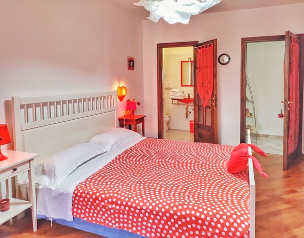 Un dormitorio con cama, mesita de noche con lámpara, colcha roja con lunares y decoración con forma de corazón rojo.