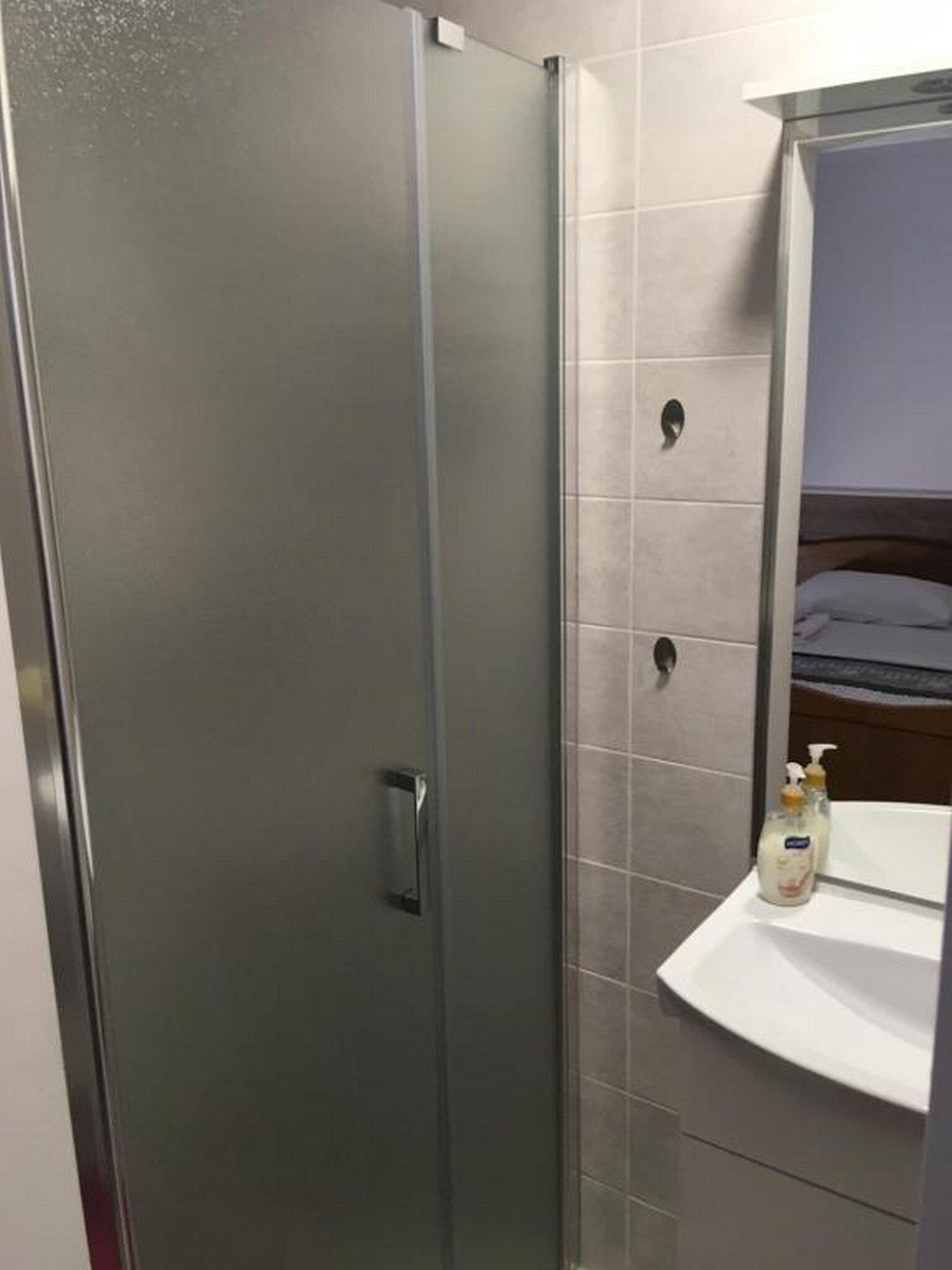 A bathroom with a glass shower door, a white sink with a mirror above it, and a bottle of hand soap on the sink.