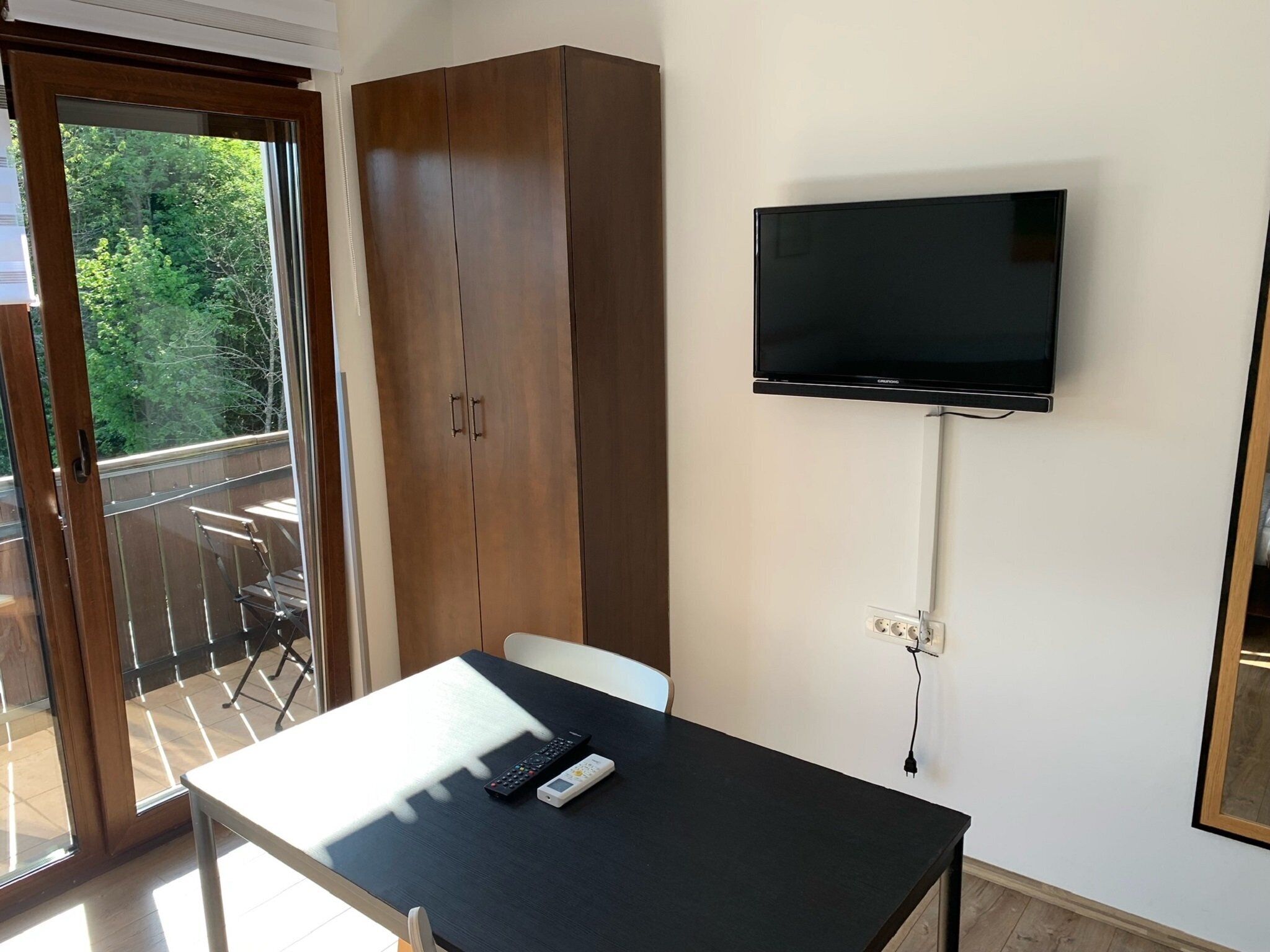 A room with a large wardrobe, a table with a remote control, and a mounted TV.
