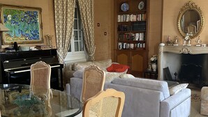 A living room with a glass coffee table, two wicker chairs, a grey sofa, a bookshelf, a piano, and a fireplace.