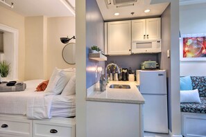 Cozy bedroom with a bed, bedside table, and lamp. Compact kitchenette with microwave, refrigerator, and sink. Seating area with a cushion for relaxation.