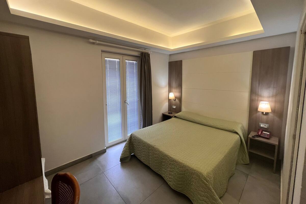 A hotel room with a bed, a chair, a desk with a lamp, and a window with blinds.