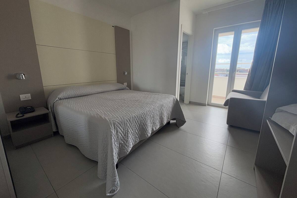 A modern hotel room with a large bed, a bedside table with a phone, a chair, and a view of the outdoors through a large window.