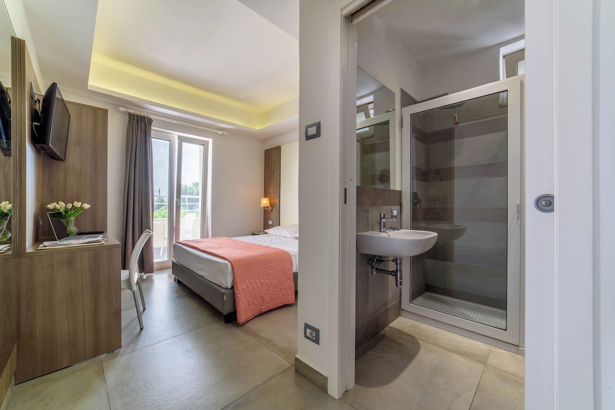 A hotel room with a bed, a desk, a chair, a shower, and a sink.