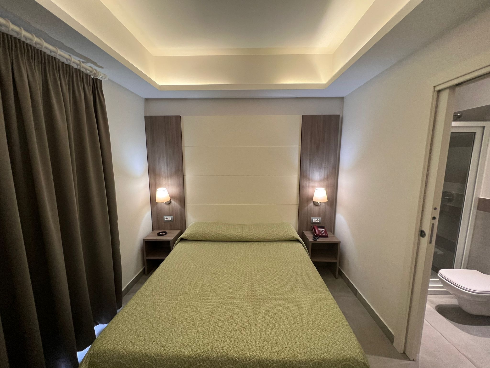 Habitación de hotel con una cama individual, dos lámparas de noche, un teléfono y una puerta que da a un baño.