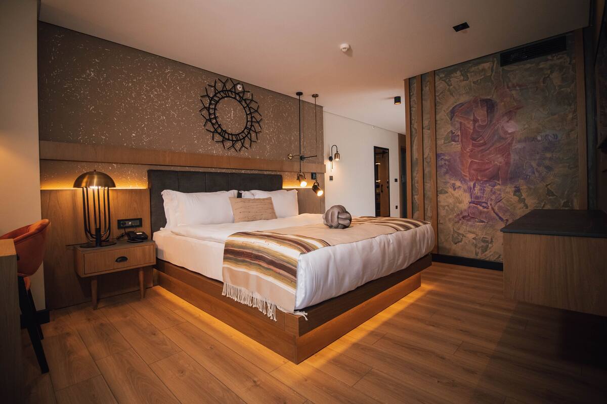 A modern bedroom with a wooden floor, a large bed with a headboard, bedside lamps, and a wall-mounted artwork.