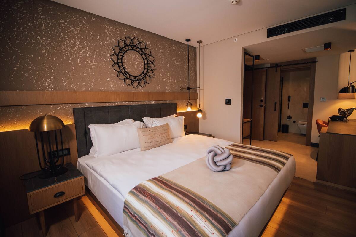A modern hotel room with a large bed, a nightstand with a lamp, a wall-mounted decorative piece, and a bathroom visible through an open door.