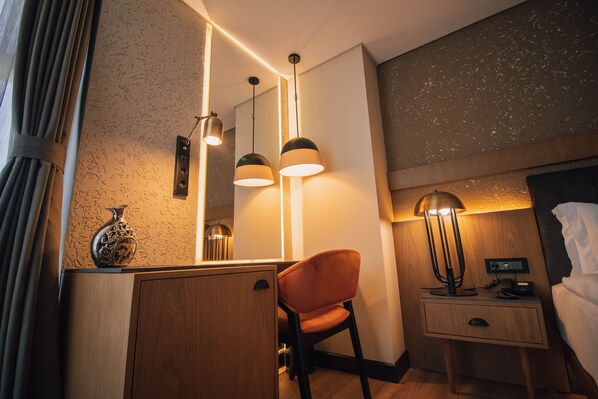 A hotel room with a wooden desk, a chair, a bedside table with a lamp, a nightstand, and a large mirror. - Panagia Premier Trabzon (Trabzon)