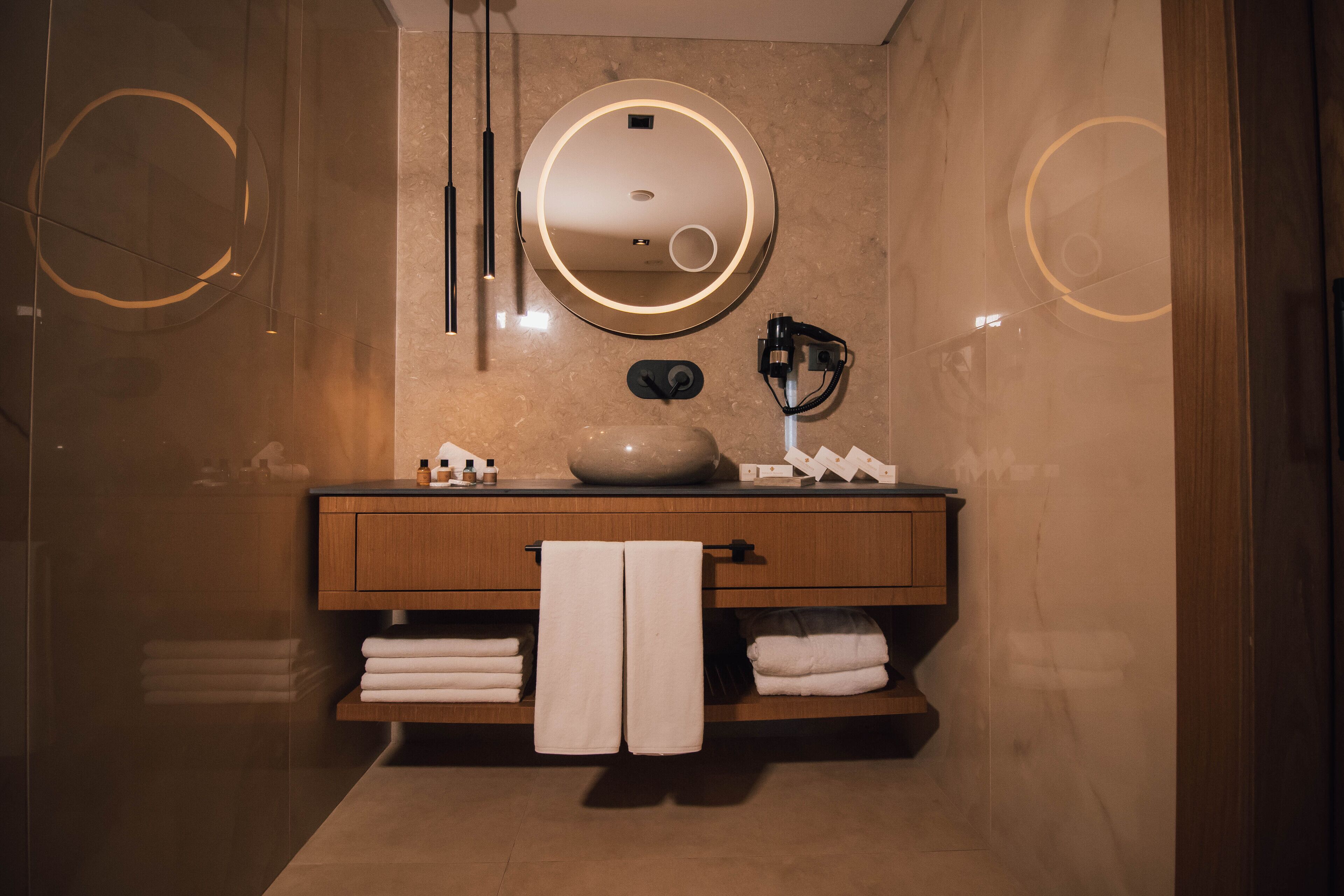 Un baño moderno con un espejo redondo, tocador de madera y toallas ordenadamente dobladas.