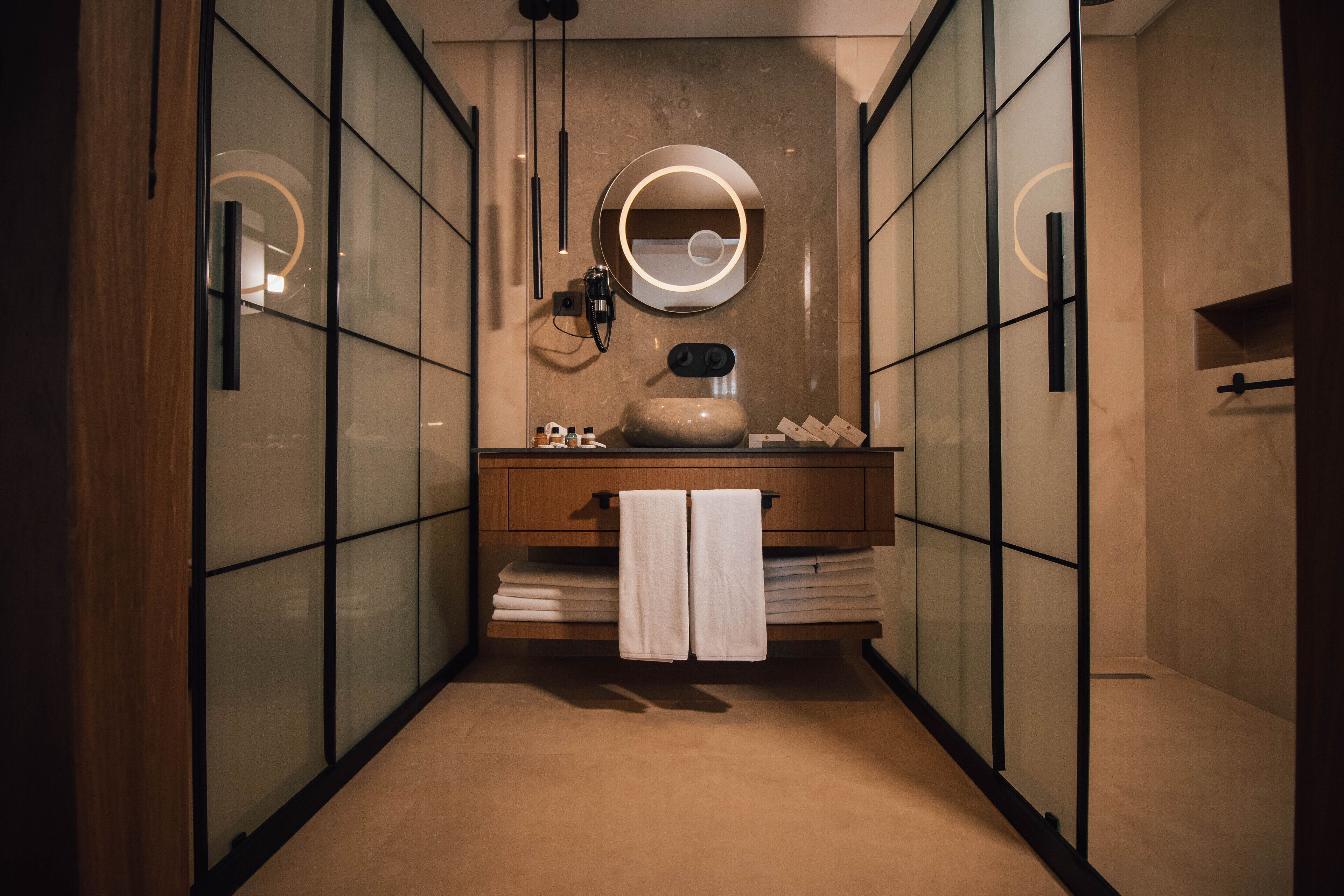 Een moderne badkamer met een glazen douchewand, een houten wastafelmeubel, een ronde spiegel en een handdoekhouder.