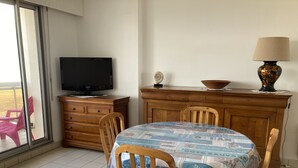 A dining area with a table covered in a nautical-themed tablecloth, wooden chairs, a wooden cabinet with a television, and a window with a view of outdoor furniture. - Apartment (Saint-Hilaire-de-Riez)