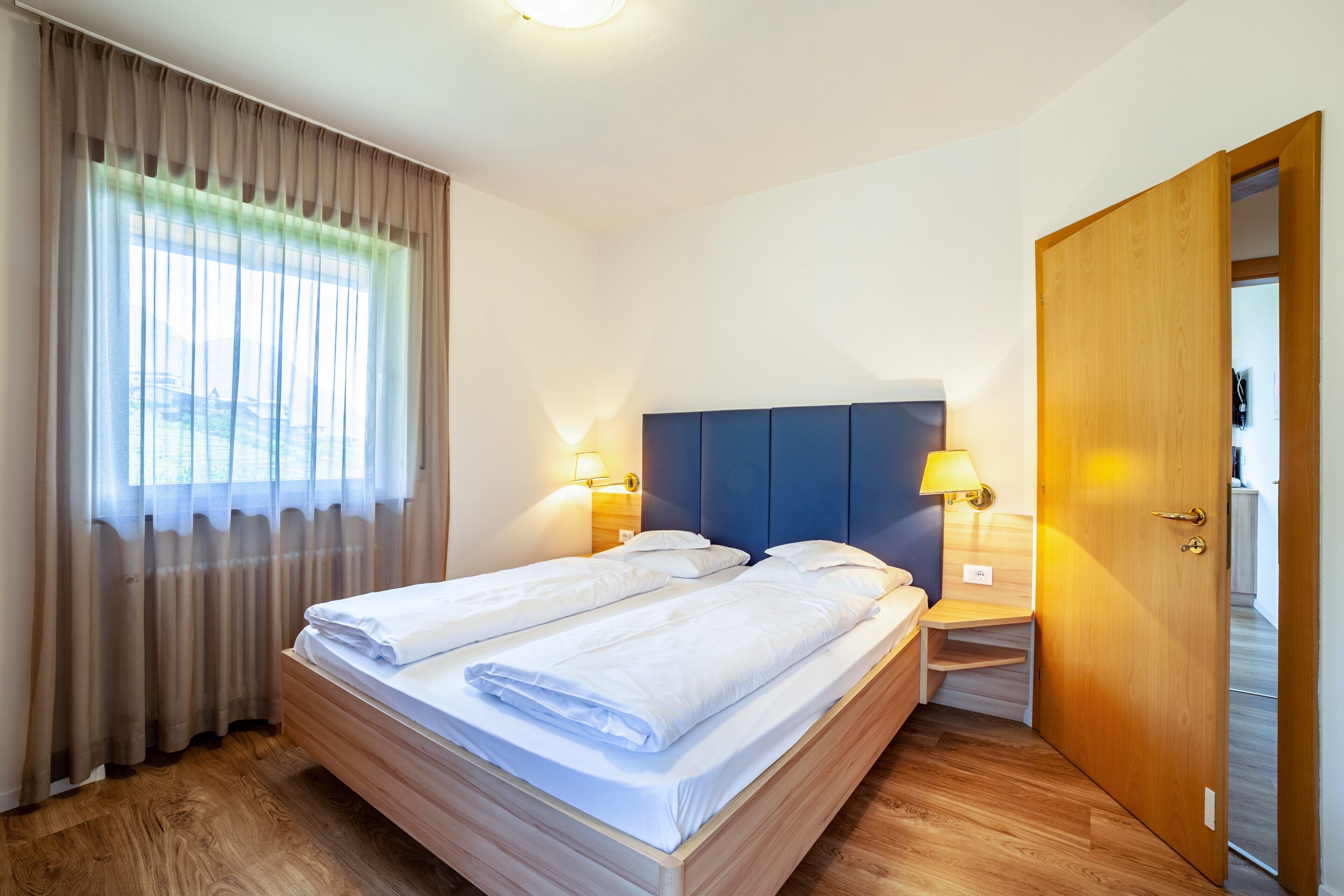 Una cama doble con cabecera de madera, dos mesitas de noche con lámparas, una puerta de madera y una ventana con cortinas.