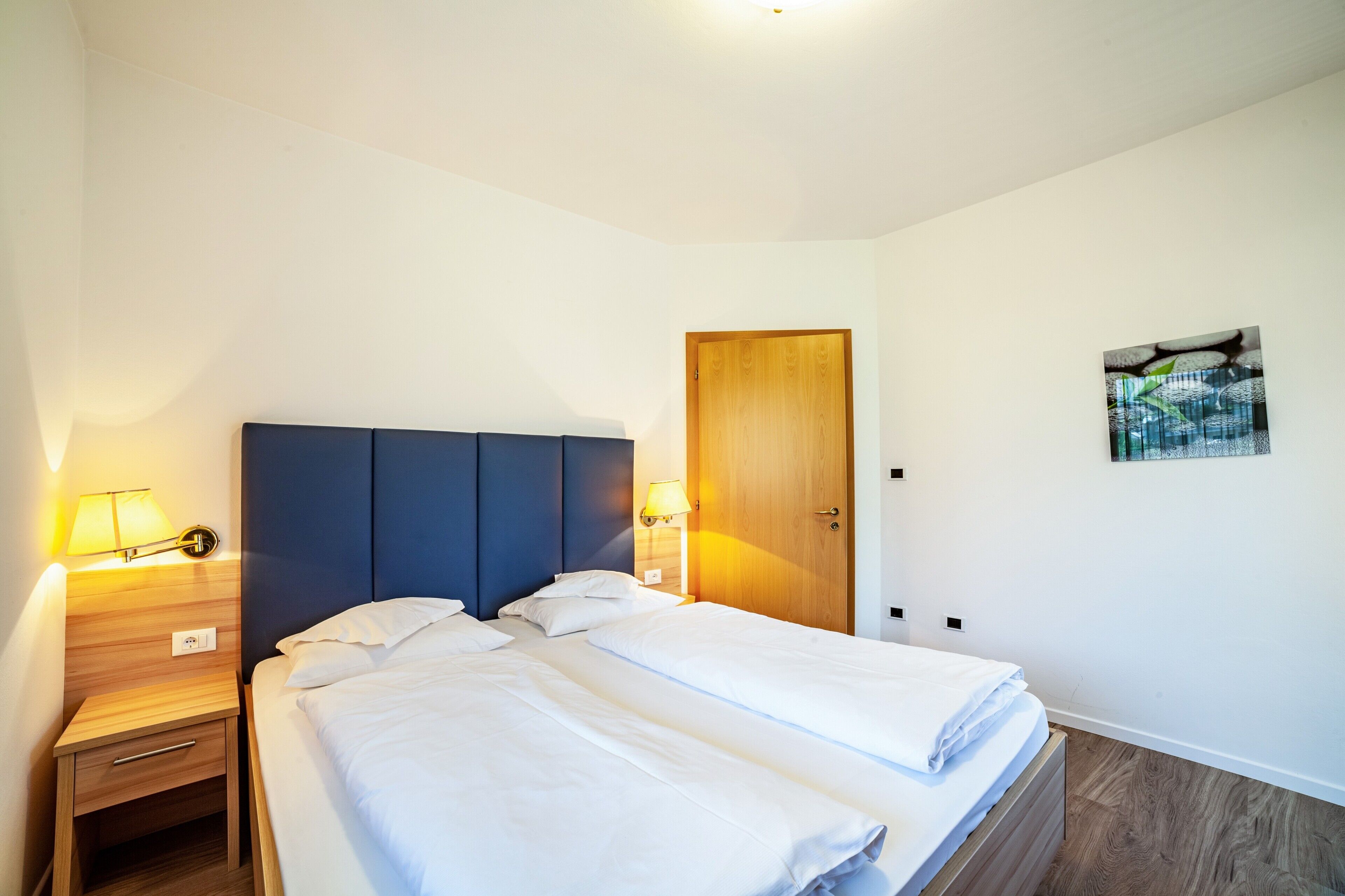 Een tweepersoonsbed met witte lakens, een houten nachtkastje met een lamp, een houten deur en een kunstwerk aan de muur.